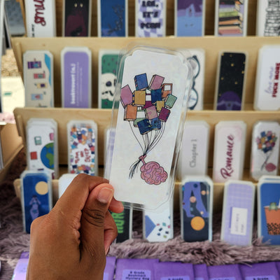 Hand holding a bookmark with a brain holding balloons shaped like books.